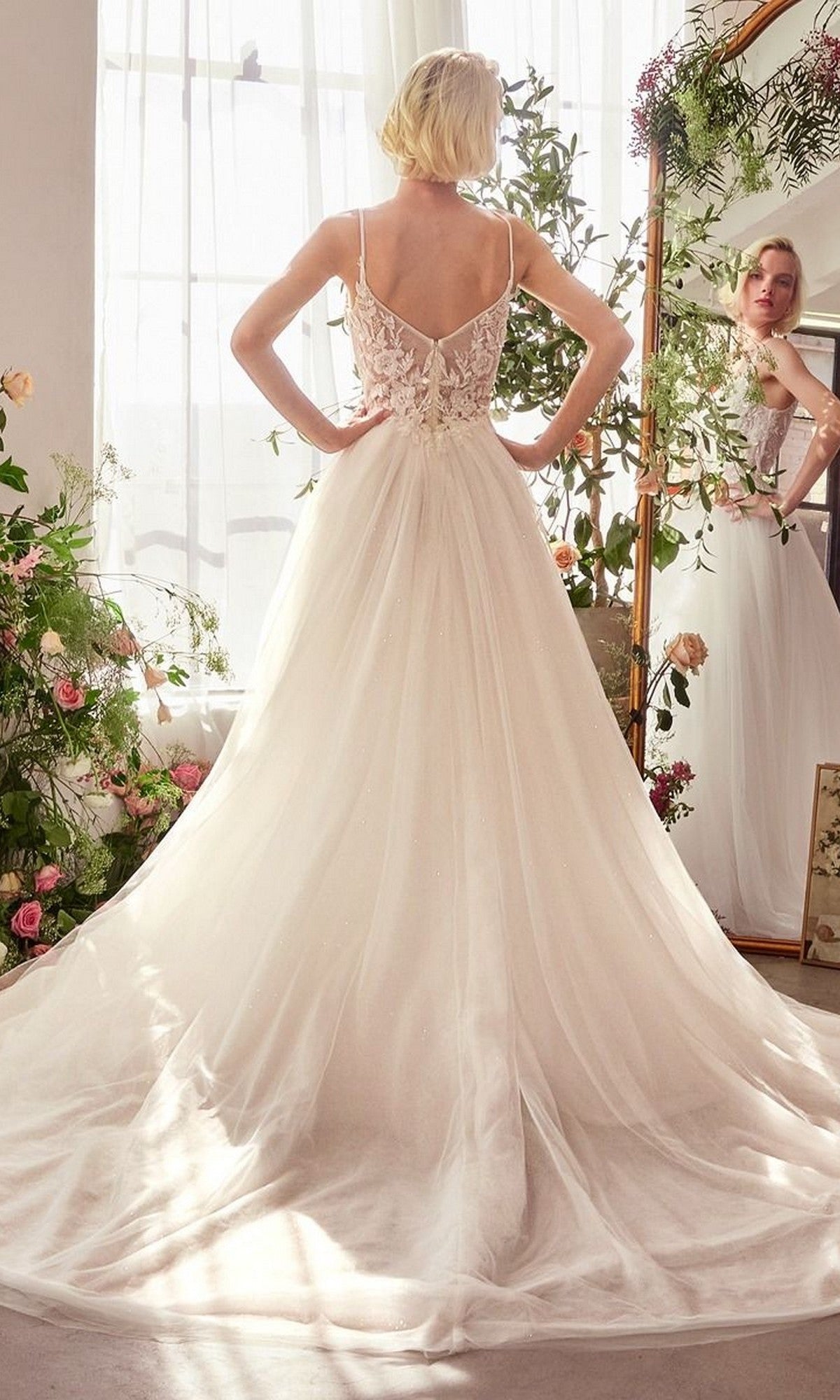A woman wearing the Andrea and Leo WL029 long wedding dress with a sleeveless lace corset bodice and flowing tulle A-line skirt stands facing away, gazing into a flower-framed mirror lit by natural light.